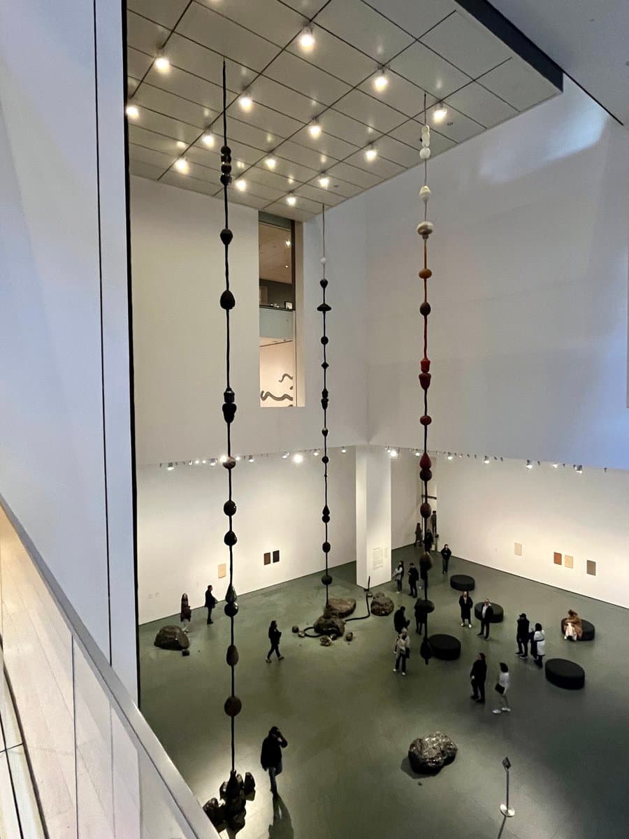 A high-angle photograph of a spacious, white-walled museum gallery featuring a large-scale art installation with long vertical strings of beads and stones suspended from the ceiling, along with rocks on the floor and visitors walking through the space.