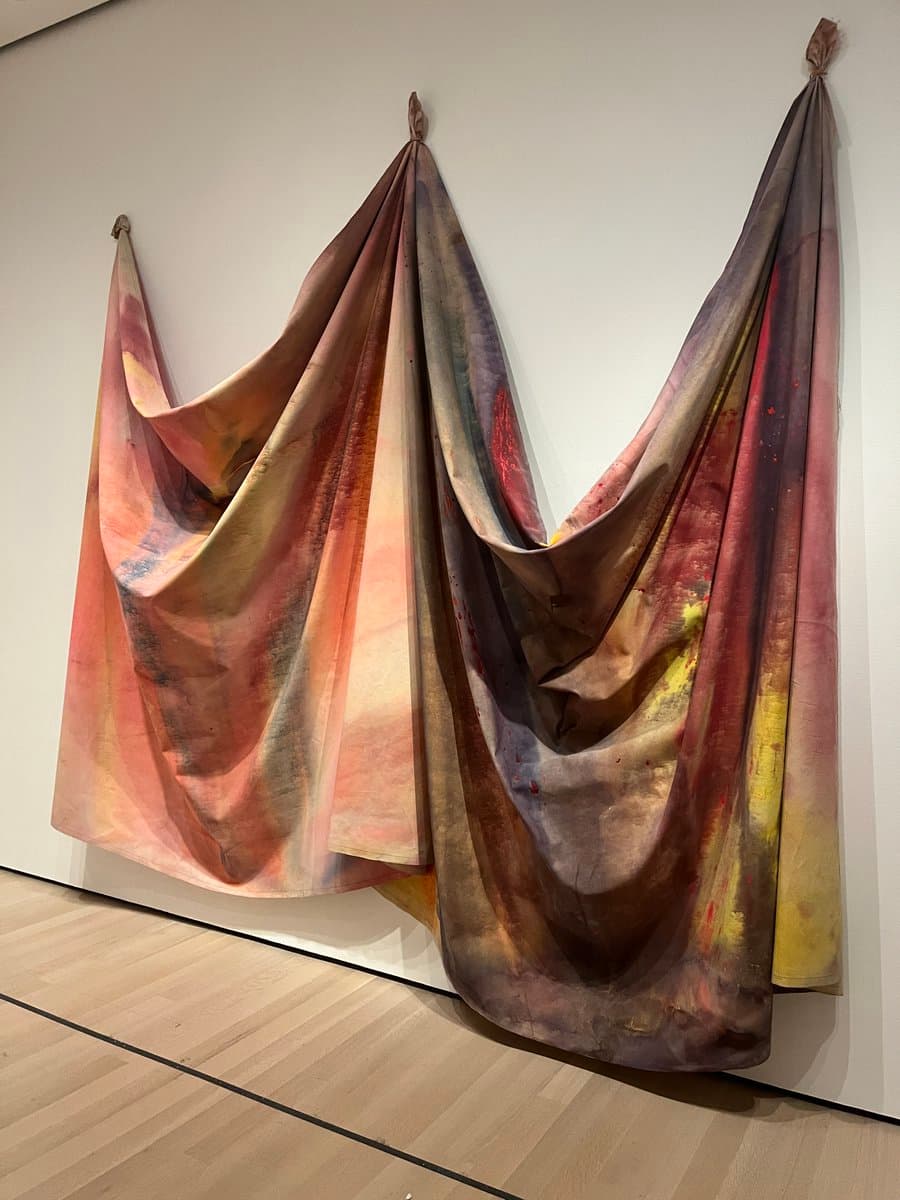 A photograph of a large, draped textile art installation hanging from a white gallery wall, featuring soft folds in shades of pink, purple, and yellow.