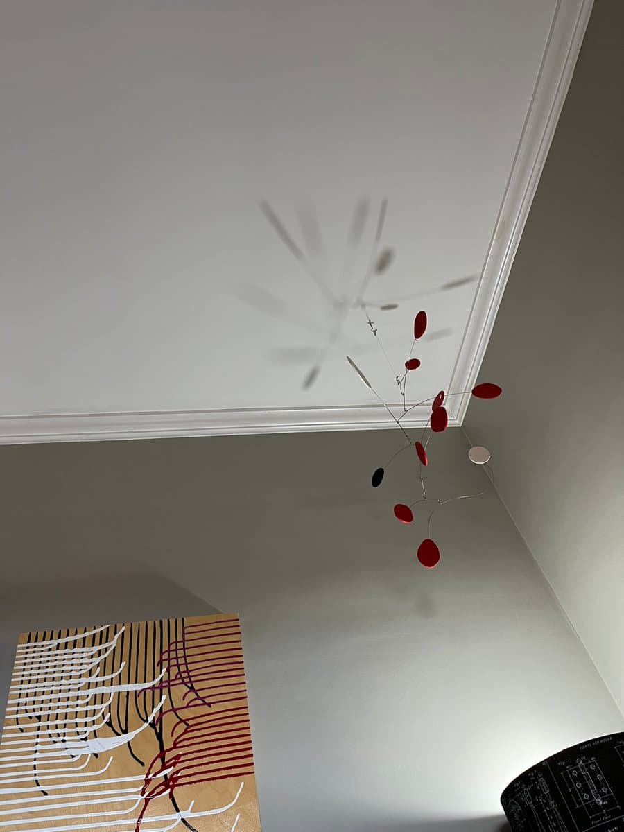 A mobile sculpture with red and black oval shapes hangs from the corner of a white ceiling, casting a blurred shadow on the wall above a piece of abstract art.