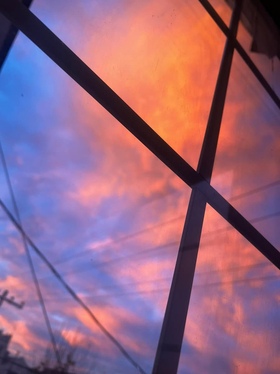 A view of a colorful sunset sky with orange and blue clouds, seen through a window with dark diagonal bars and power lines in the foreground.