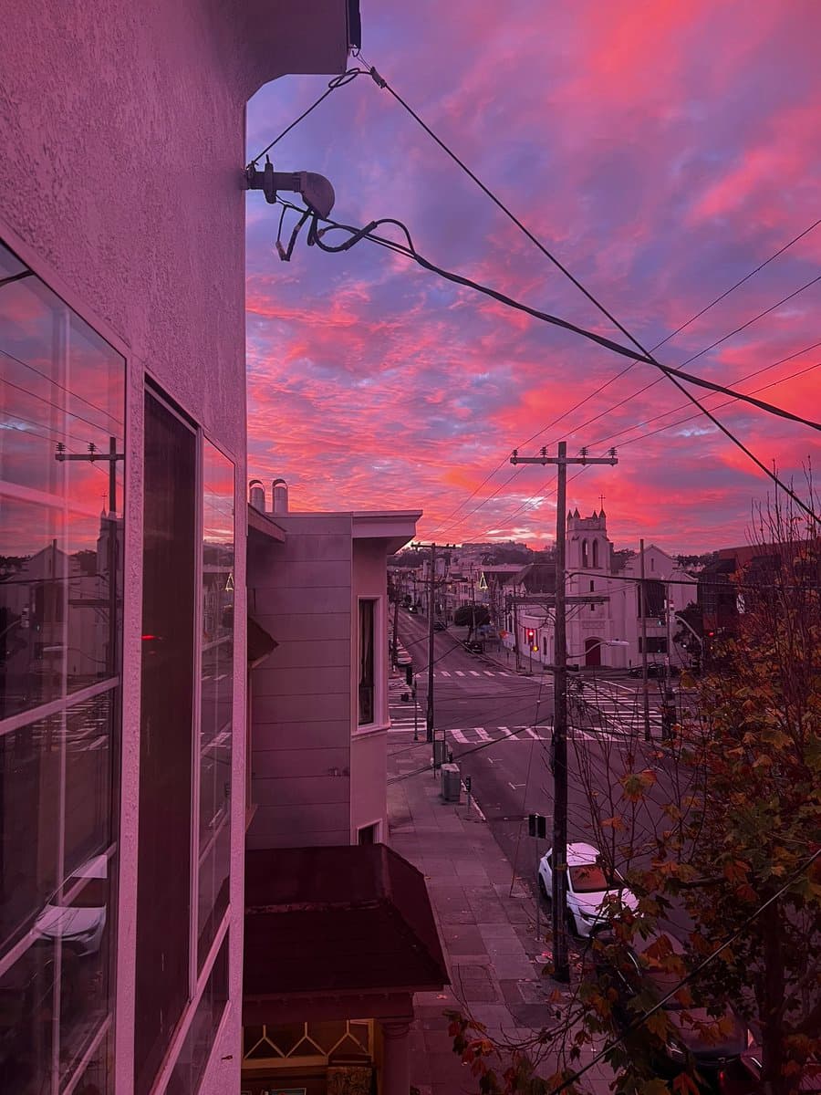 A photograph taken from an elevated vantage point overlooking a city street at sunset, featuring a dramatic sky filled with pink and purple clouds. The scene includes a building facade on the left, a street with a crosswalk and parked cars below, and a white church with a steeple in the distance.