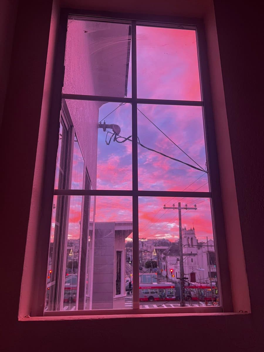 A view through a six-paned window looking out onto a city street at dusk, with the sky illuminated in vibrant shades of pink and purple. The scene includes a white church, a red bus, and power lines, all bathed in the warm light of the sunset.