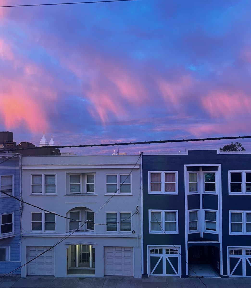 A photograph of a row of multi-story townhouses, painted in white and dark blue, set against a dramatic sky with pink and purple sunset clouds. Overhead power lines stretch across the foreground.