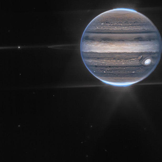 A high-resolution image of the planet Jupiter against a black, star-filled background, showing its distinct horizontal cloud bands and the Great Red Spot. The planet is partially illuminated with a bright blue atmospheric glow along its edge, and horizontal lens flare streaks extend from the left side of the frame.