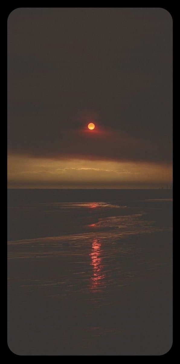 A vertical photograph of a sunset over a body of water, featuring a small, bright orange sun low in a dark, cloudy sky. The sun's reflection creates a shimmering red path on the rippling water surface below.