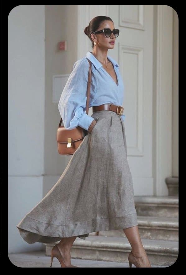 A woman wearing a light blue button-down shirt, a long beige skirt, and sunglasses walks down a set of stone steps. She carries a brown leather shoulder bag and wears a brown belt with a gold buckle.