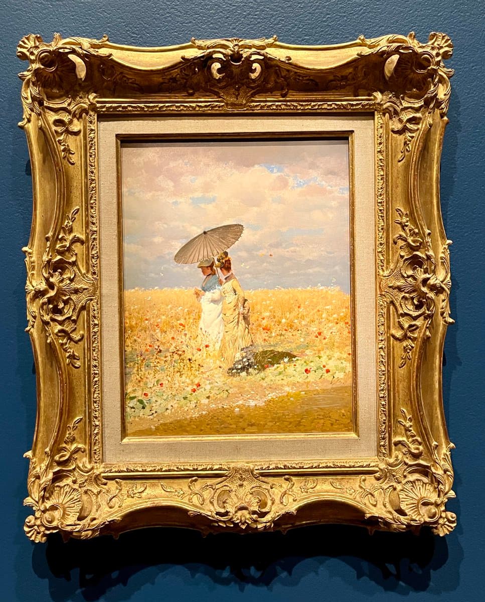A photograph of a framed Impressionist painting hanging on a dark blue gallery wall. The artwork depicts two women in a field of tall grass and wildflowers, with one holding a parasol over the other. The painting is enclosed in an ornate, gilded frame with elaborate floral and scrollwork detailing.