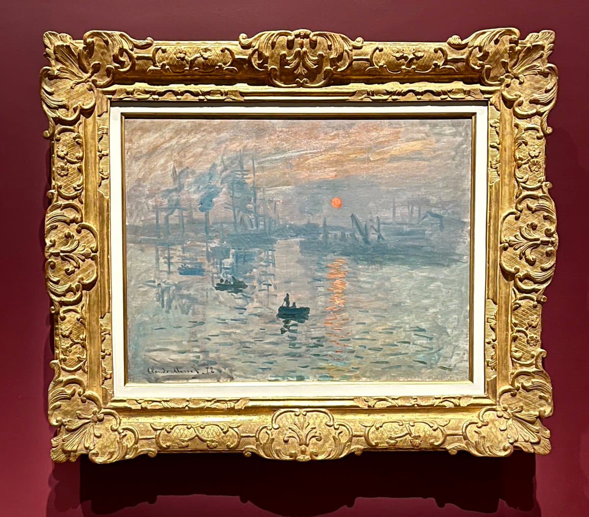 A photograph of a framed Impressionist painting depicting a harbor scene with a setting sun, displayed on a dark red gallery wall. The artwork is enclosed in an ornate, gilded frame with intricate floral and scrollwork detailing.