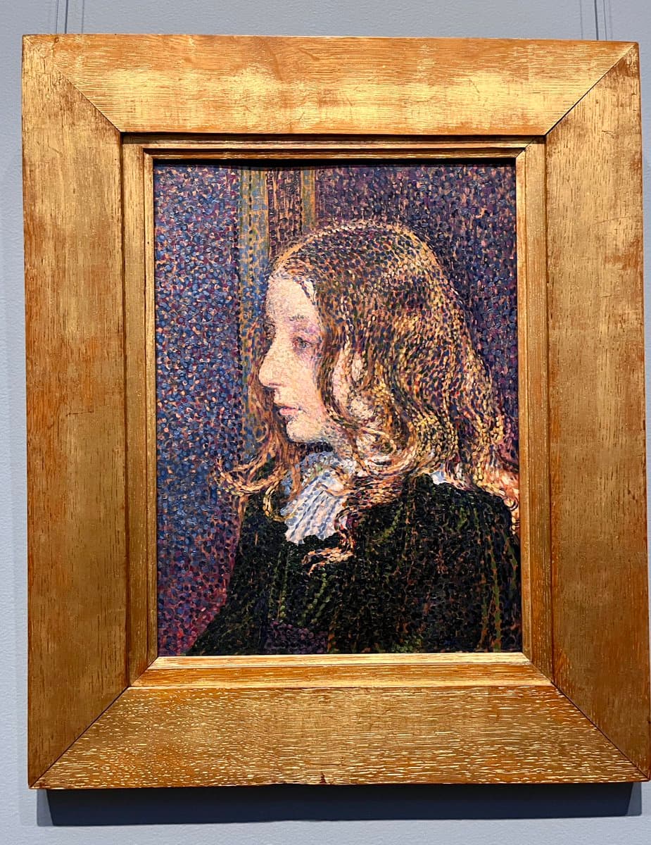 A framed painting in a gallery, depicting a woman with long, wavy hair in profile. The artwork is executed in a pointillist style, using small, distinct dots of color to form the image.