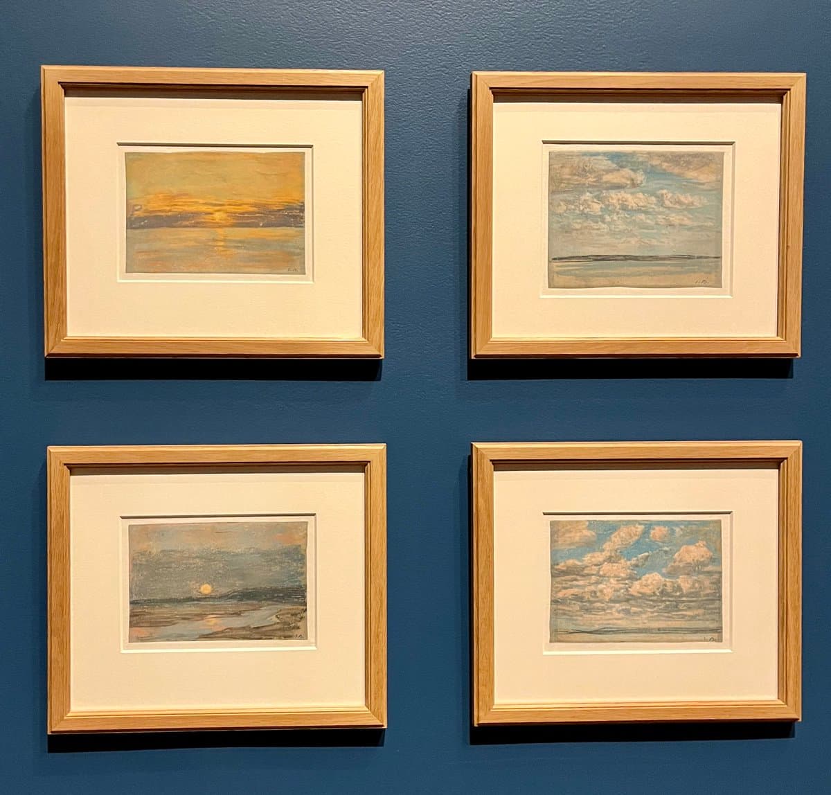 Four framed Impressionist landscape paintings are displayed in a two-by-two grid on a dark blue gallery wall. The artworks feature soft, textured brushstrokes depicting skies, clouds, and water in warm and cool tones.