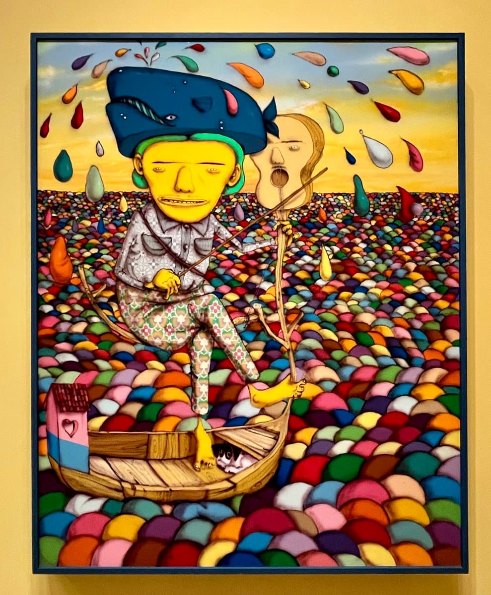 A framed, colorful painting by OSGEMEOS depicting a surreal scene of a figure with a blue whale for a hat sitting in a boat on a sea of multicolored pebbles. The artwork is displayed on a yellow gallery wall, showcasing the artists' vibrant and whimsical style.