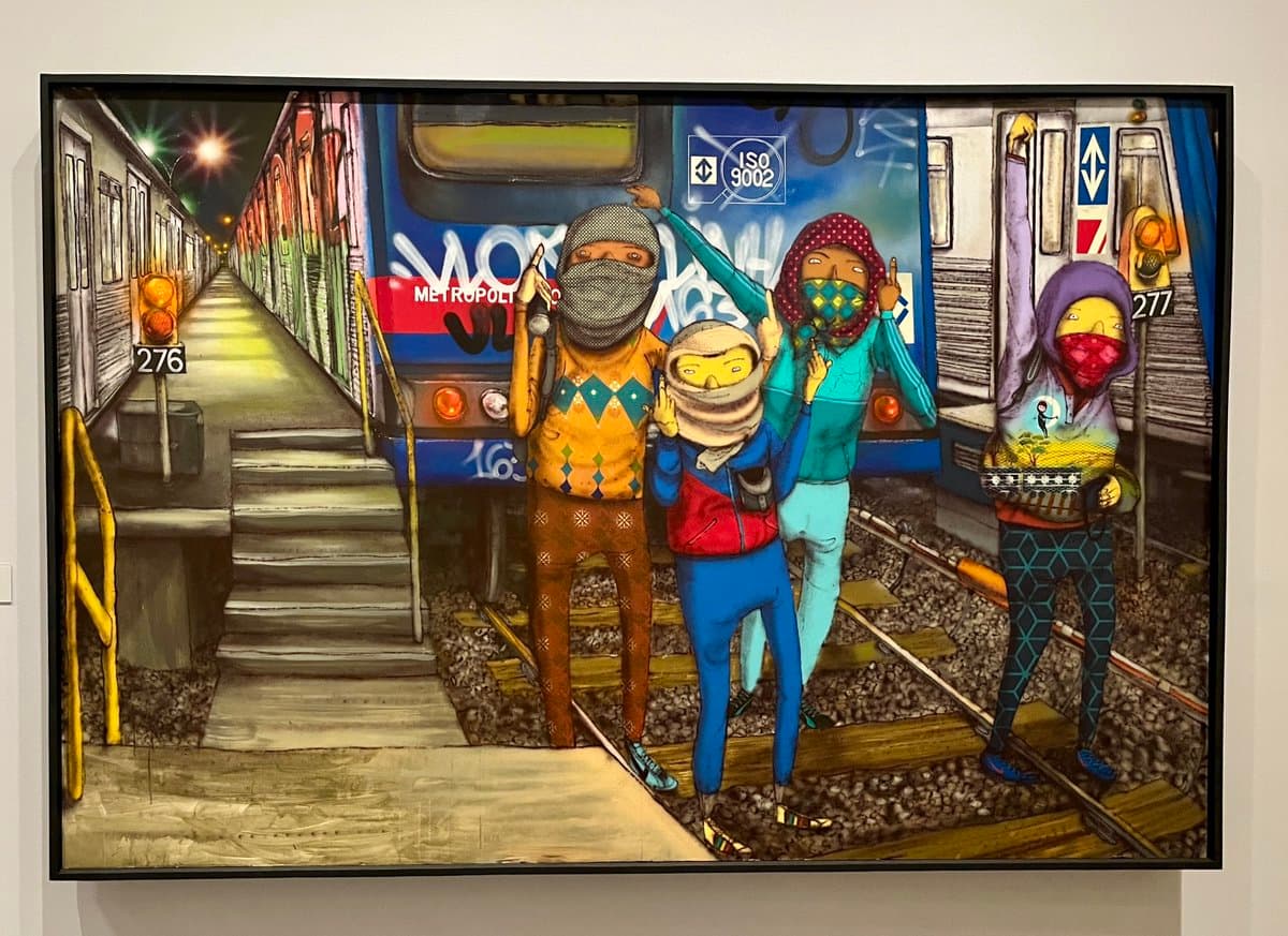 A framed, colorful painting depicting four masked figures posing on a subway track in front of a train. The artwork is displayed on a white gallery wall.