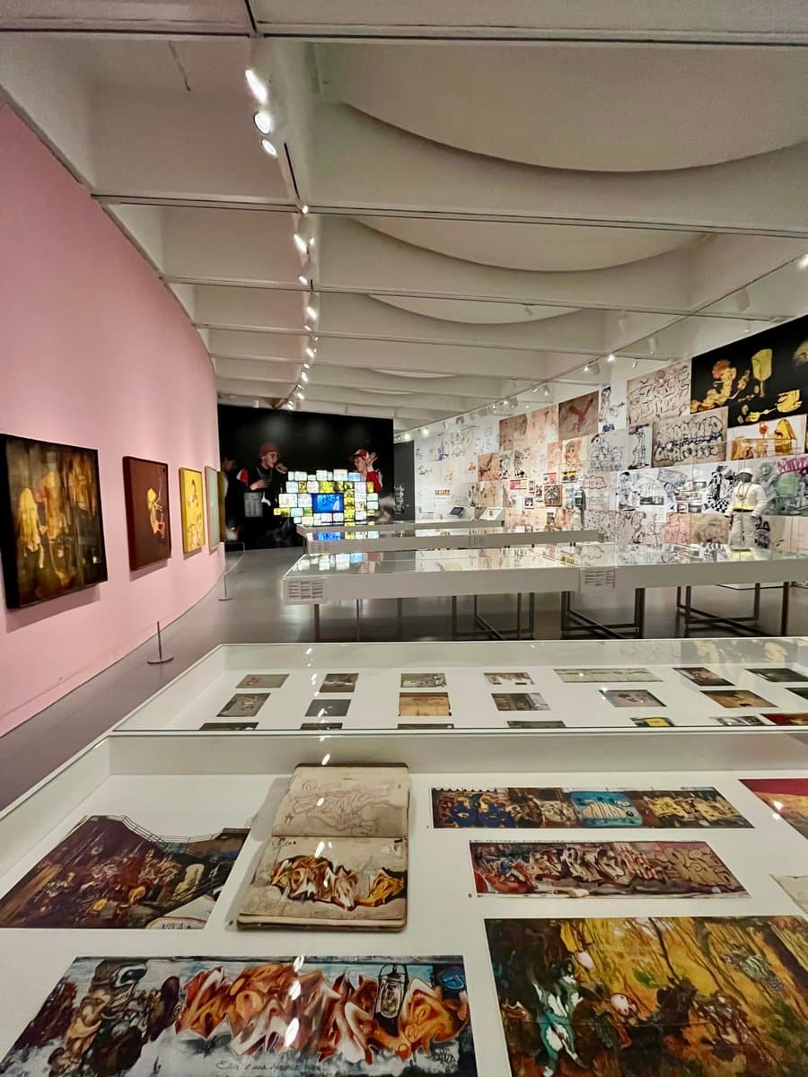 A wide-angle photograph of a brightly lit art gallery featuring a colorful OSGEMEOS exhibit. The space includes a pink wall with framed paintings on the left, glass display cases in the foreground containing sketchbooks and artwork, and a wall covered in graffiti-style drawings on the right.