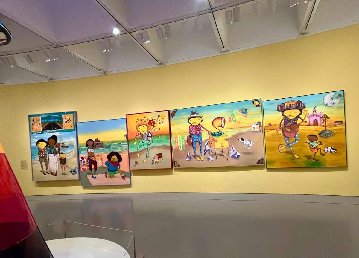 A wide shot of a gallery wall displaying five colorful, cartoon-style paintings by the artist OSGEMEOS, featuring stylized figures in various outdoor settings. The artworks are mounted on a yellow wall beneath track lighting in a museum or exhibition space.