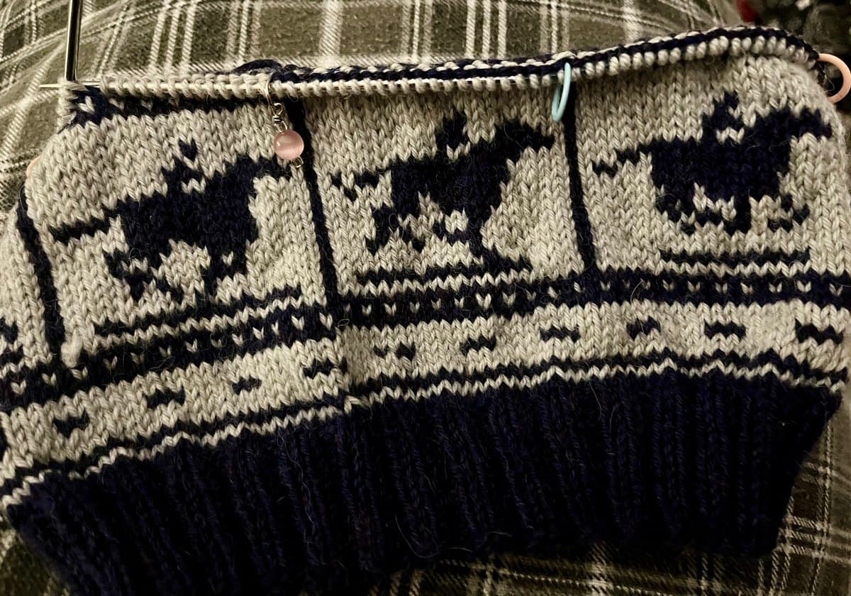 A close-up photograph of a knitting project in progress, featuring a dark blue and cream pattern with a repeating motif of a castle or building. The work is held on a knitting needle with a pink bead marker and a blue stitch marker attached.