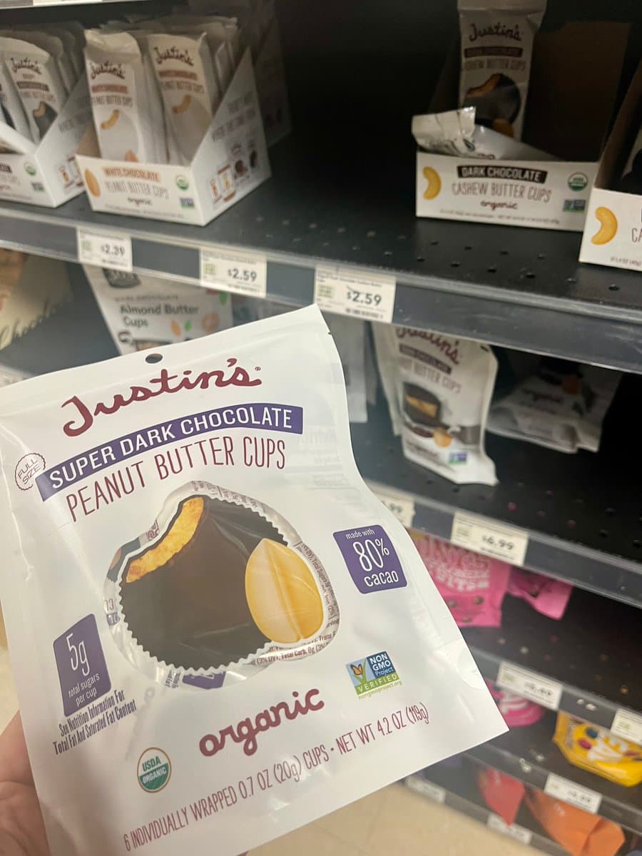A close-up photograph of a hand holding a package of Justin's Super Dark Chocolate Peanut Butter Cups in a grocery store aisle. The package is white with purple and red text, featuring an image of a chocolate-covered peanut and a label indicating it is made with 80% cacao. In the background, store shelves display other Justin's products, including white chocolate and dark chocolate varieties, along with price tags.