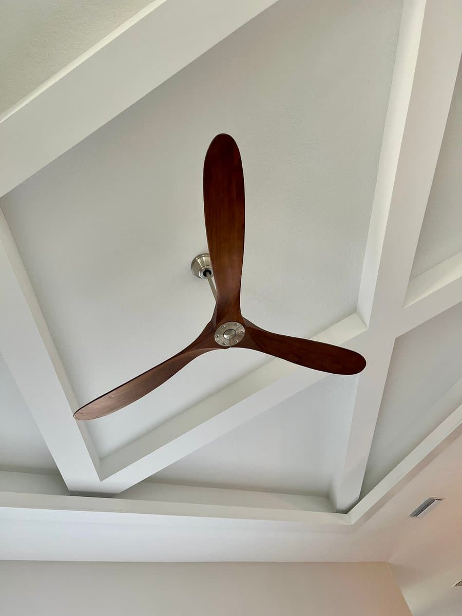 A low-angle photograph looking up at a three-bladed wooden ceiling fan mounted on a white, coffered ceiling. The fan has a metallic central hub and is attached to the ceiling via a short downrod. The surrounding ceiling features recessed rectangular panels and a small air vent is visible in the lower right corner.