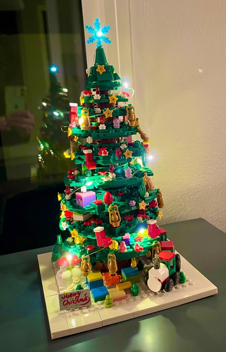 A detailed LEGO Christmas tree model decorated with lights, ornaments, and small figures, standing on a white baseplate with a small train and a 'Merry Christmas' sign.