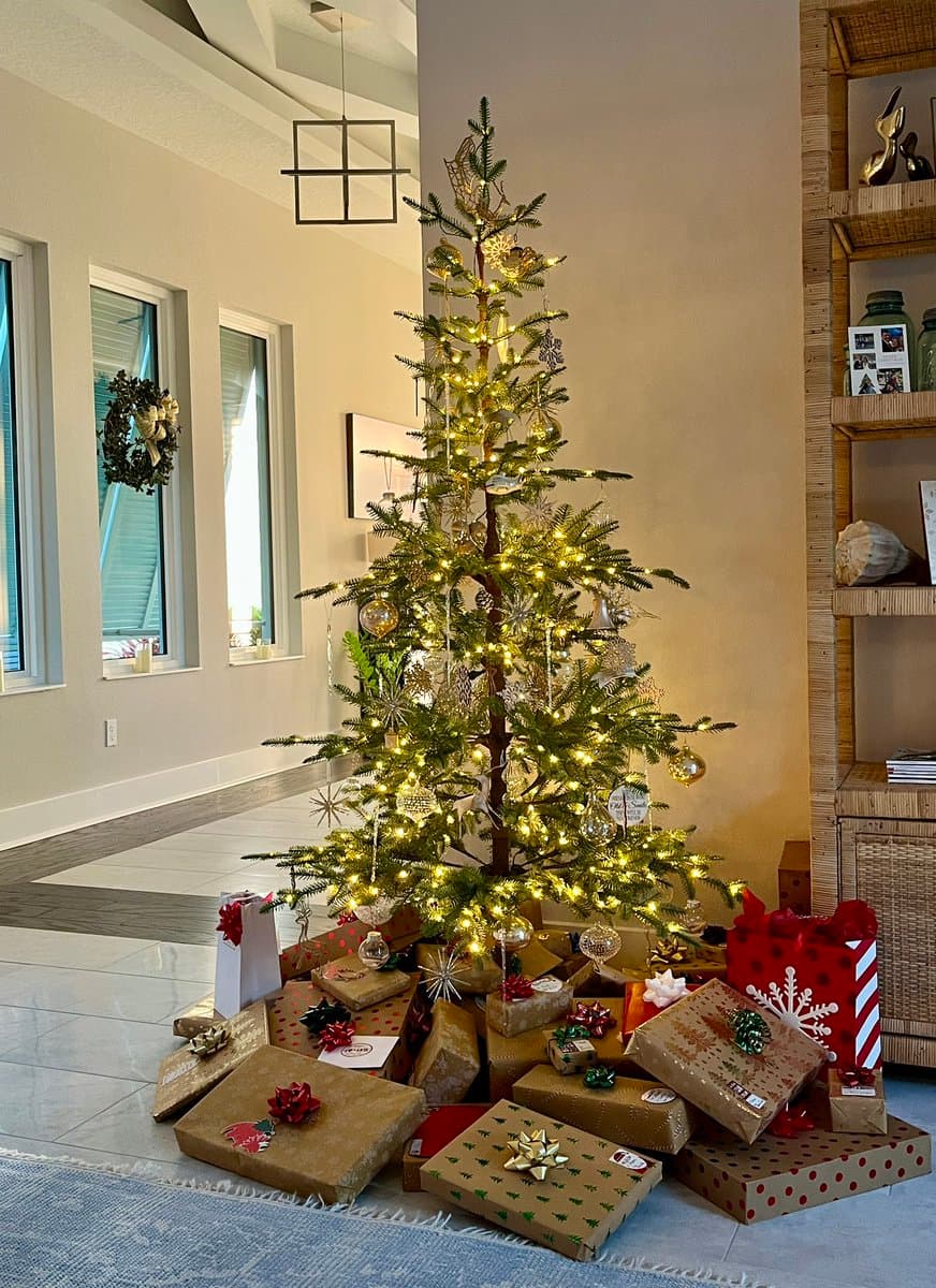 A decorated Christmas tree with warm white lights and silver ornaments stands in a living room, surrounded by numerous wrapped gifts and gift bags on the floor. The scene includes a wreath on a window and a wooden shelving unit in the background.