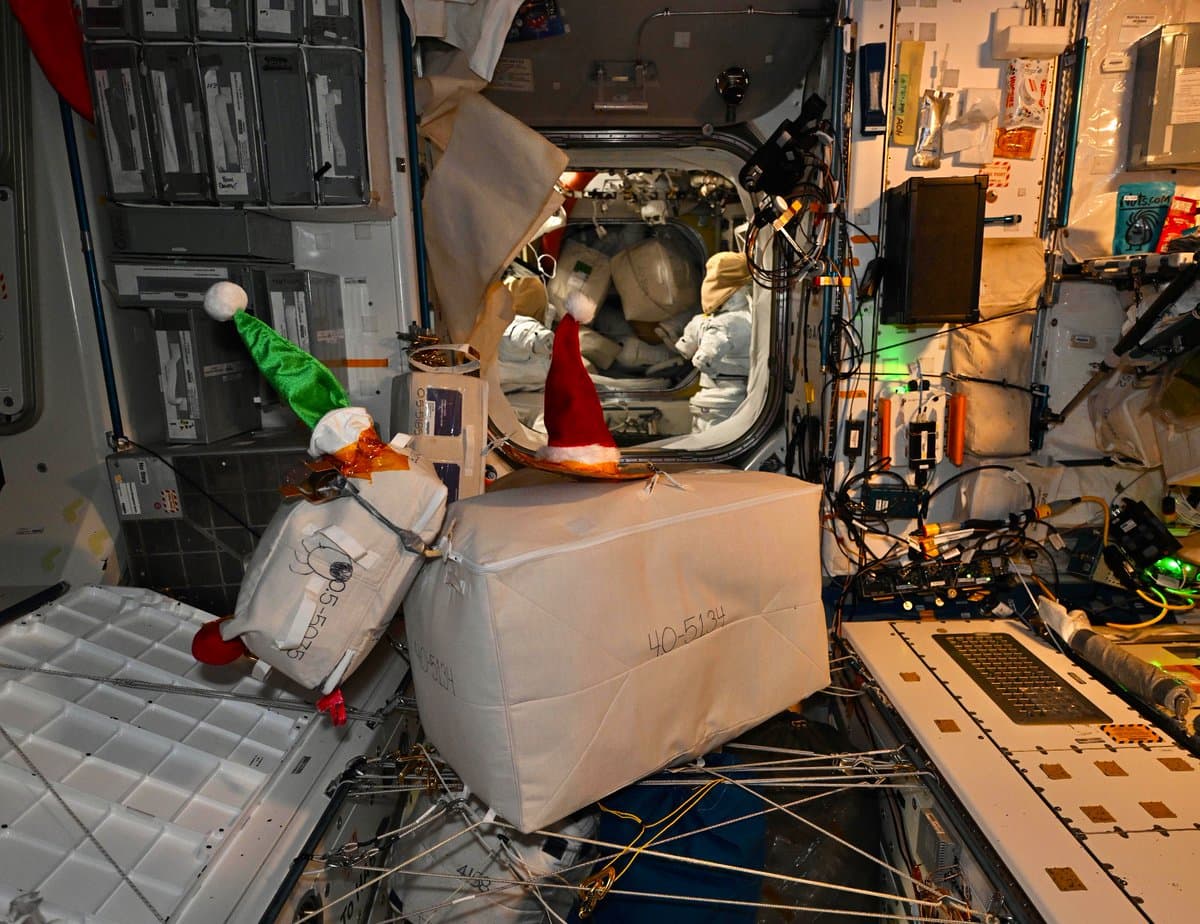 A photograph taken inside a space station module showing white cargo bags decorated with red and green Santa hats. The background reveals the interior of the station, including equipment, cables, and a person in a white spacesuit visible through a hatchway.