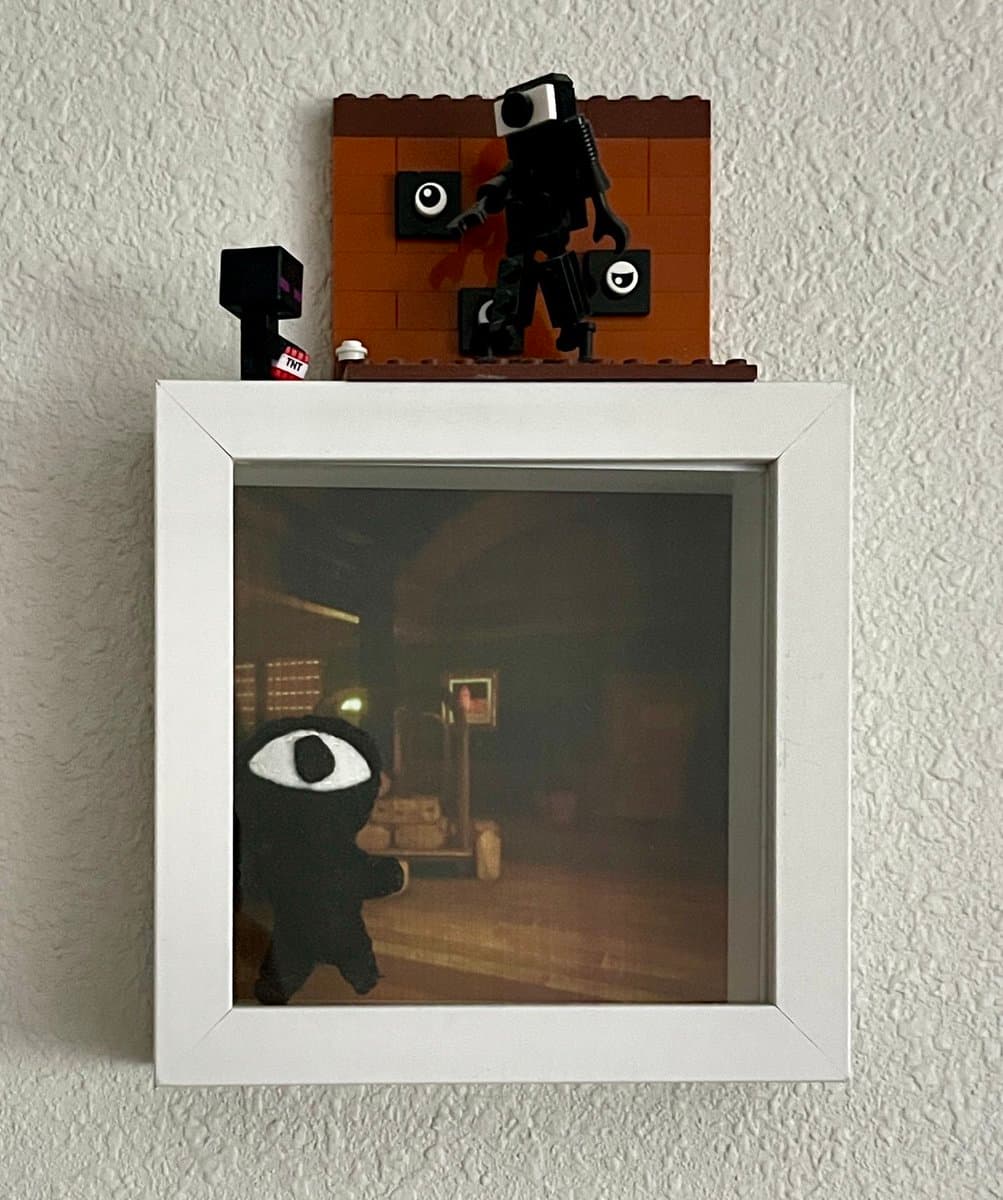 A photograph of a white picture frame on a wall, containing a screenshot of a dark, first-person video game scene featuring a black, eye-headed character. A small LEGO diorama of a similar character is mounted on top of the frame.