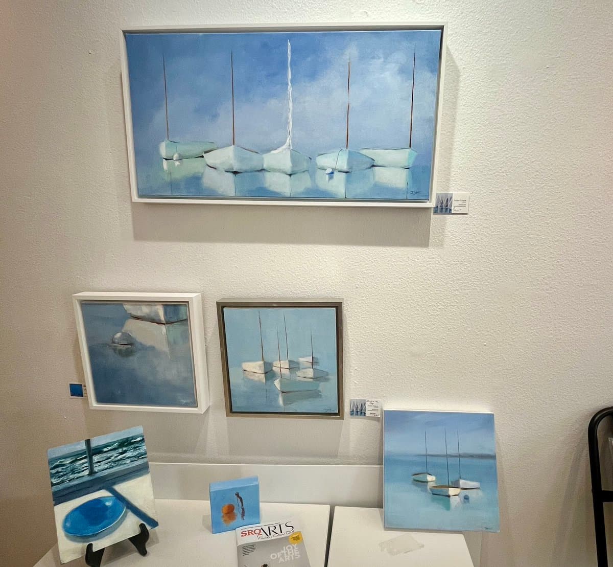 A display of several framed and unframed paintings in an art gallery, featuring a consistent theme of boats and water in blue and white tones. The artworks are arranged on a white wall and on a table below, with small informational placards next to each piece.