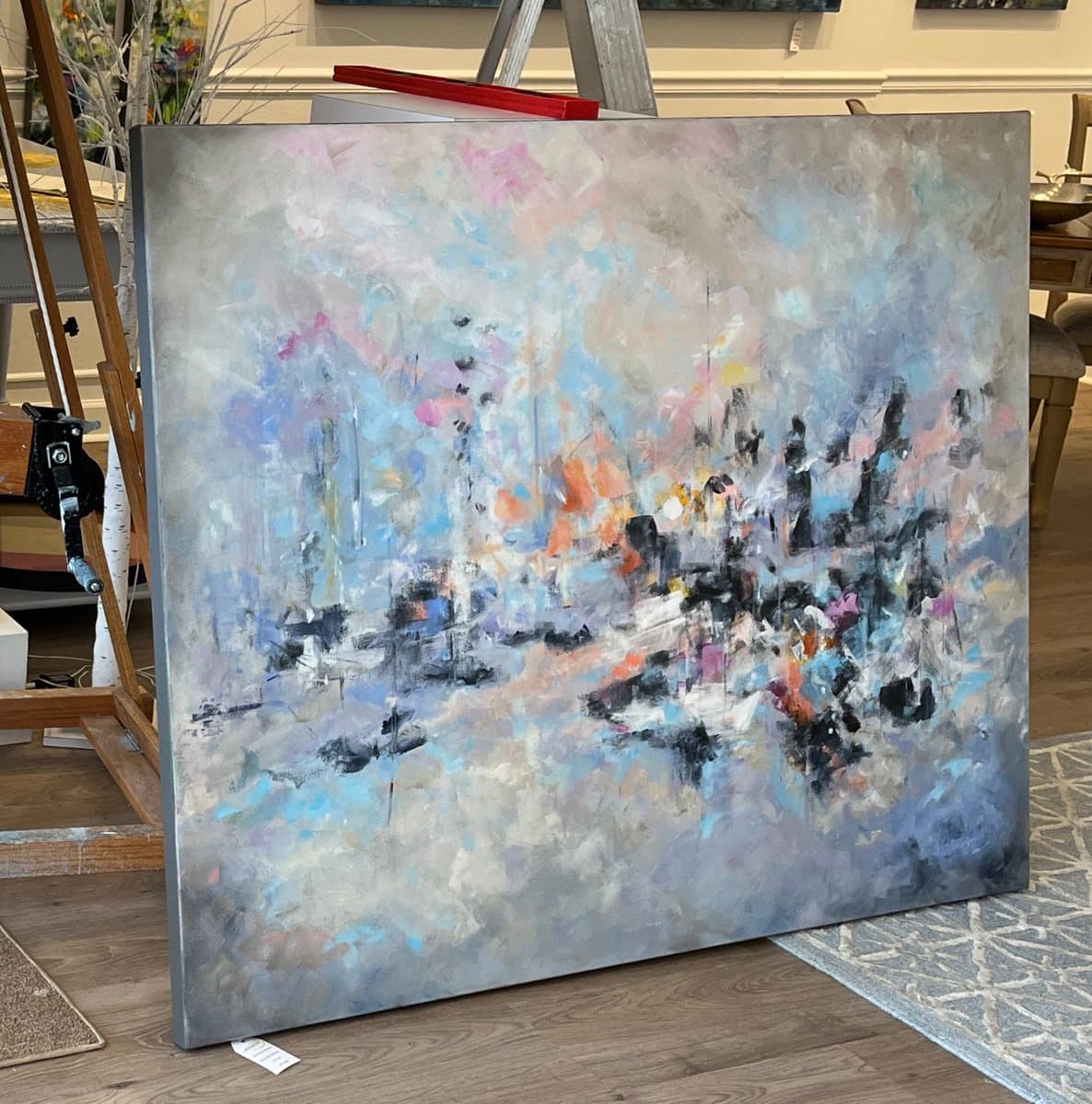 A large, square abstract painting featuring soft, blended strokes of grey, blue, and pink with darker accents, leaning against a wooden easel in an art gallery setting.
