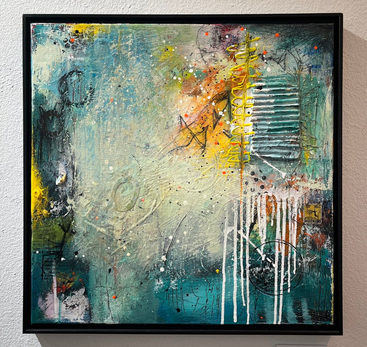 A framed abstract painting hangs on a white gallery wall, featuring a textured composition of teal, white, and yellow paint with splatters and drips. The artwork includes a prominent yellow spiral element and vertical white streaks.
