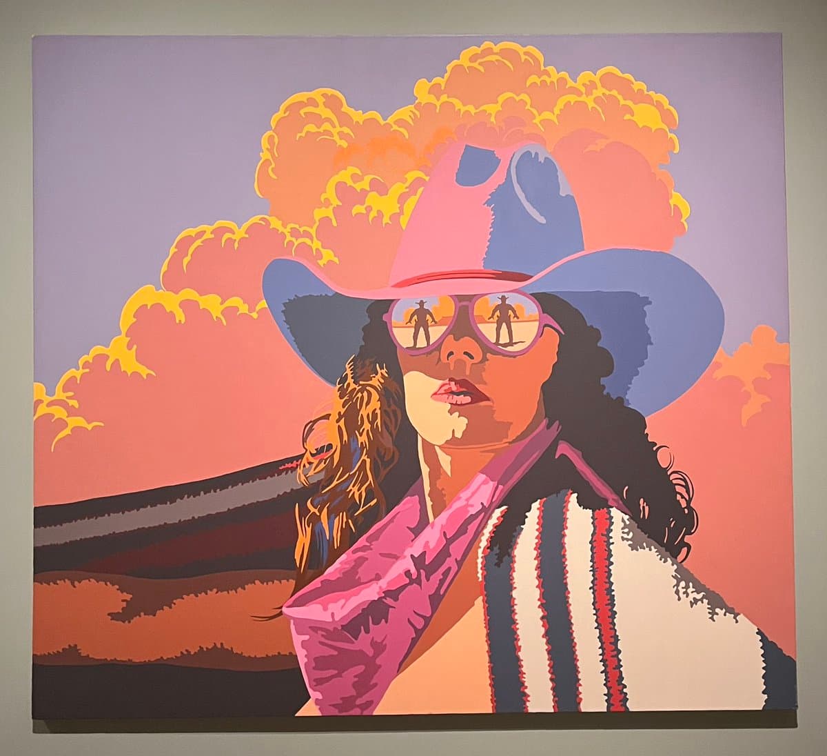 A photograph of a colorful, stylized painting depicting a person with dark curly hair wearing a pink cowboy hat and sunglasses. The figure is shown from the chest up against a backdrop of a pink and purple sky with large, orange clouds.