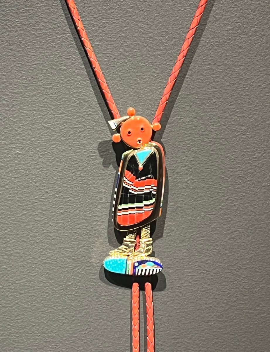A close-up photograph of a colorful, stylized figurine pendant hanging from a braided orange cord against a textured gray background. The figure has an orange head, a black and orange patterned body, and is wearing blue and white sneakers.