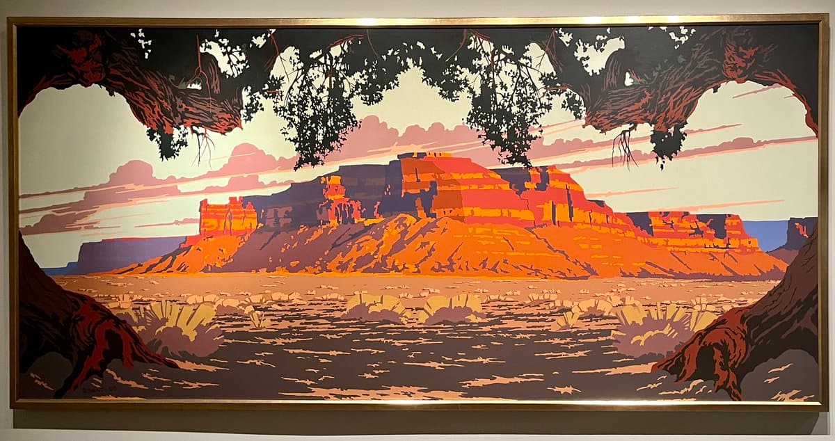 A framed, panoramic artwork in a stylized, flat-color illustration style depicting a desert landscape with a large mesa at sunset, viewed through the branches of trees in the foreground.