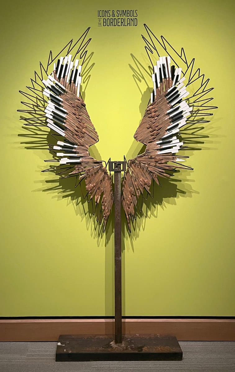 A museum sculpture of a pair of wings made from sharpened pencils and wire, mounted on a metal stand against a green wall. The piece is titled 'Icons & Symbols of the Borderland.'