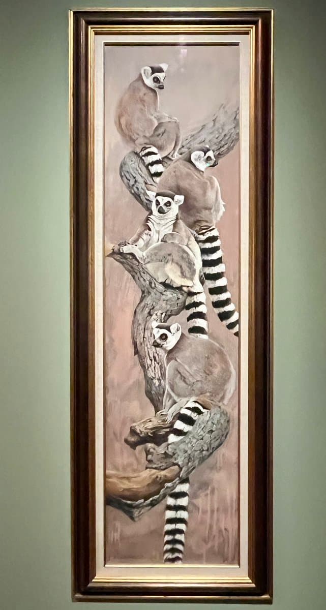 A framed painting depicting a group of ring-tailed lemurs perched on a tree branch. The artwork is displayed in a tall, vertical frame with a gold and dark wood finish, hanging on a plain wall.