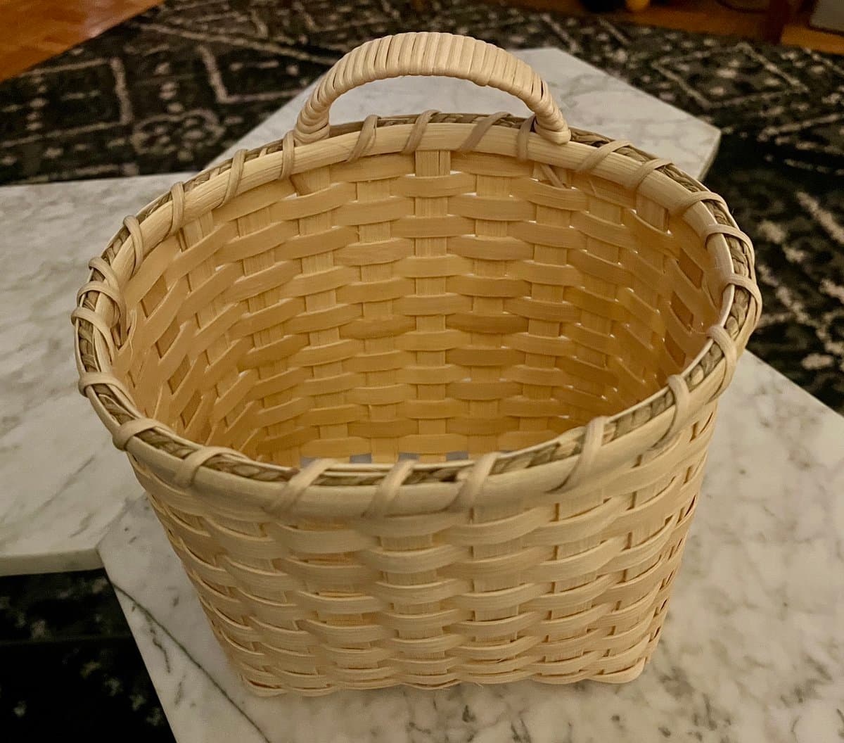 A close-up photograph of a newly woven, light-colored basket with a single handle, sitting on a white marble surface.