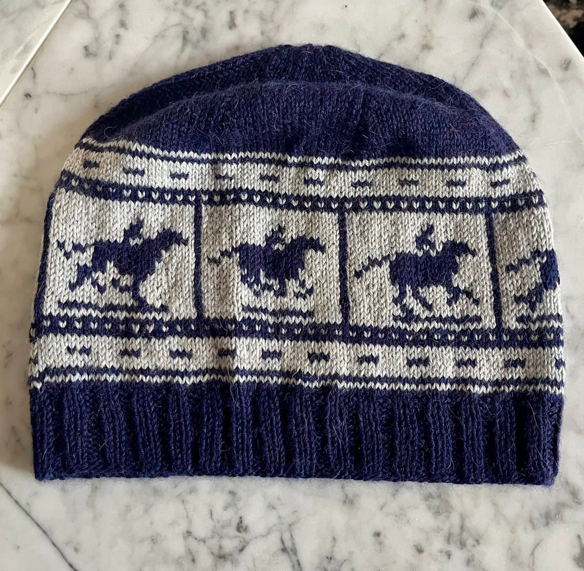 A top-down view of a hand-knitted beanie featuring a navy blue and white colorwork pattern of galloping horses. The hat is laid flat on a white marble surface.