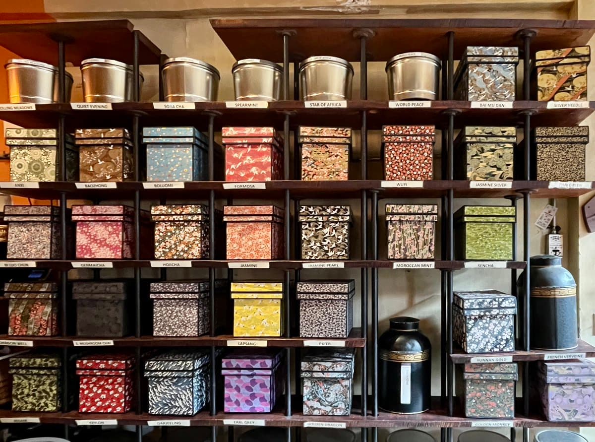 A display of numerous decorative tea tins and canisters arranged on wooden shelves, each labeled with its specific tea variety. The tins feature various patterns and colors, including floral, geometric, and solid designs, alongside several large black ceramic jars.