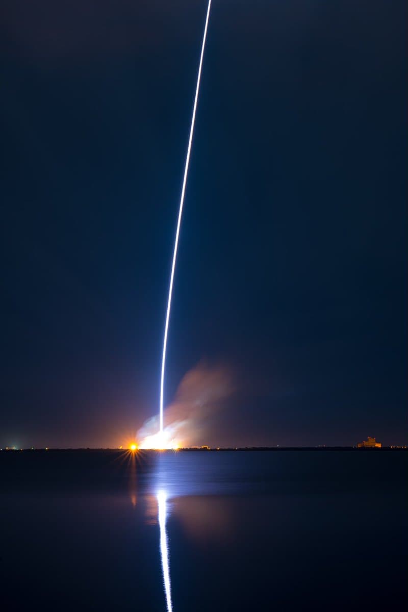 A long-exposure photograph of a rocket launch at night, showing a bright vertical light trail rising from the horizon over a body of water. The launch site glows with orange light and smoke, and the rocket's trail is reflected on the water's surface.
