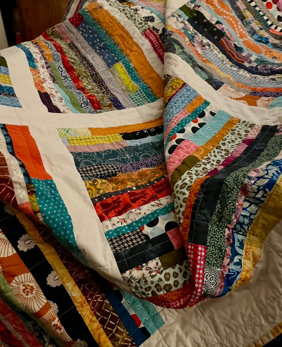 A close-up photograph of a colorful scrap quilt featuring a patchwork of various fabric strips in different patterns and colors, including floral, geometric, and animal prints, arranged in a grid-like design with white sashing.