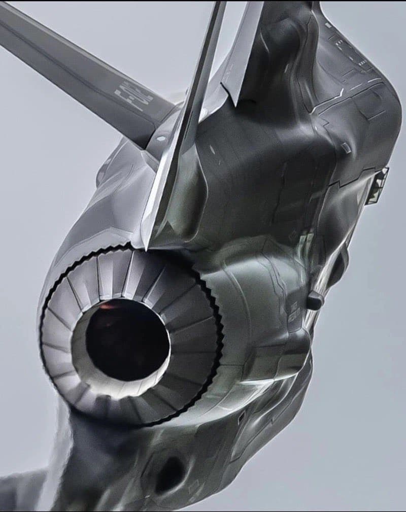 A close-up, low-angle view of a futuristic, metallic aircraft engine and fuselage against a pale sky. The image highlights the sleek, angular design of the jet's intake and surrounding body panels.