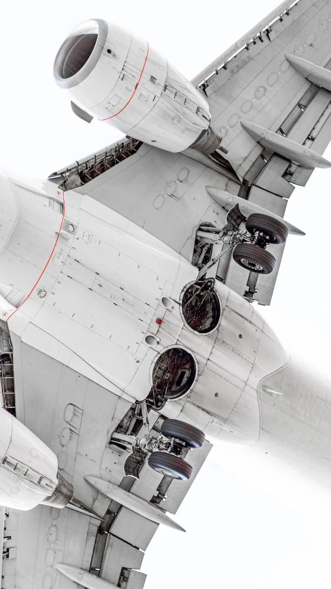 A high-angle, close-up photograph of the underside of a large white commercial airplane, showcasing its deployed landing gear, engine nacelles, and wing structure against a bright white sky.
