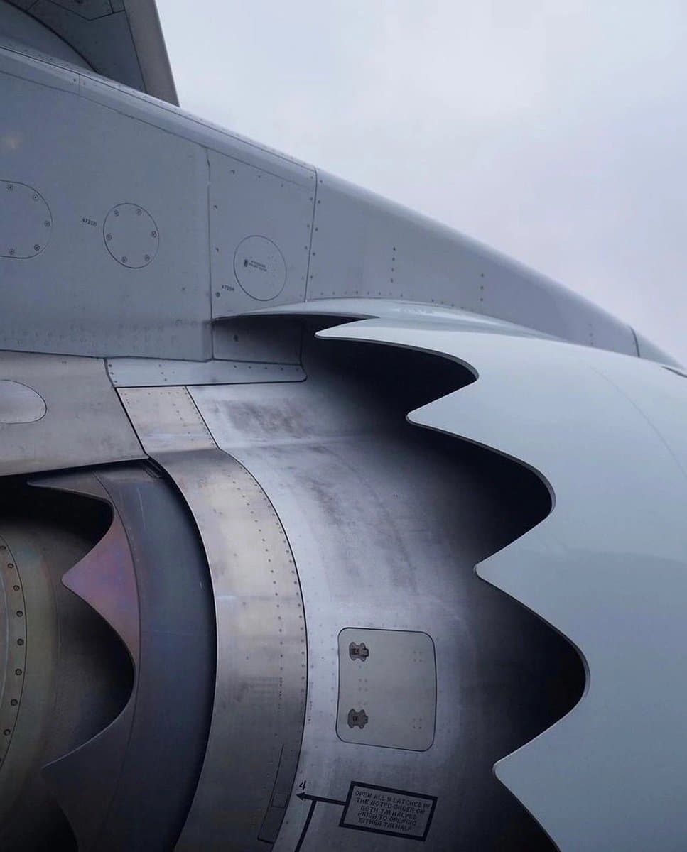 A close-up photograph of an aircraft wing and engine nacelle, highlighting the serrated, saw-toothed edge of the engine's exhaust nozzle. The image captures the metallic texture and rivets of the aerospace components against a cloudy sky.