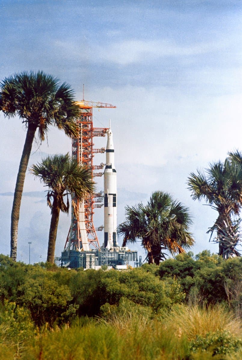 A photograph of a Saturn V rocket standing on its launch pad, surrounded by palm trees and green vegetation under a blue sky.