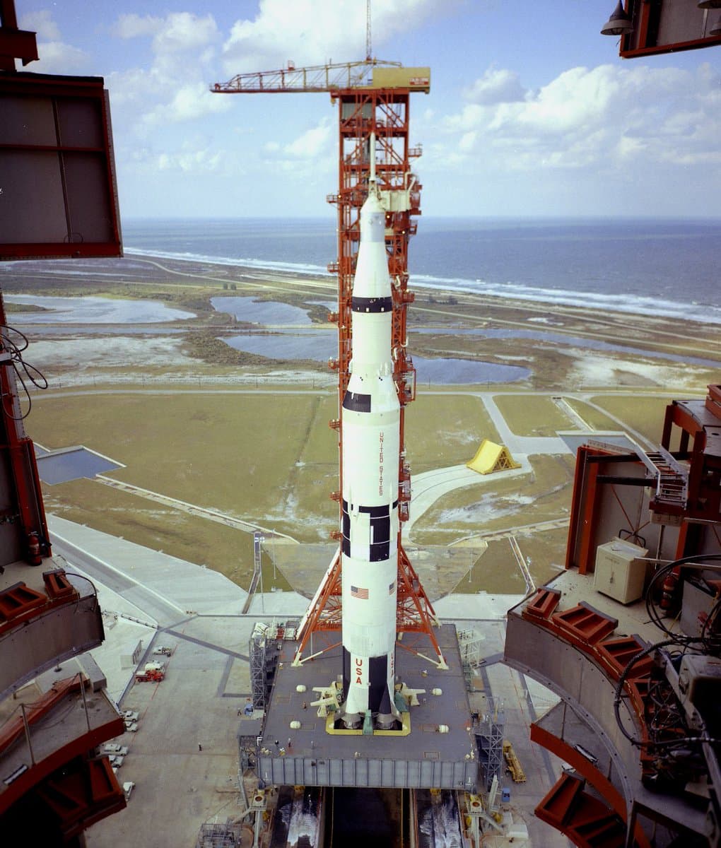 A high-angle photograph of a Saturn V rocket standing on a launch pad at a coastal space center, surrounded by launch towers and support structures.