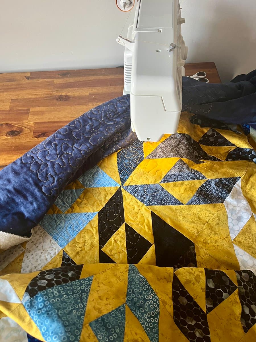 A close-up photograph of a white longarm quilting machine actively stitching a colorful patchwork quilt on a wooden table. The quilt features a geometric pattern in shades of yellow, blue, and black, with visible decorative stitching on a dark blue section.