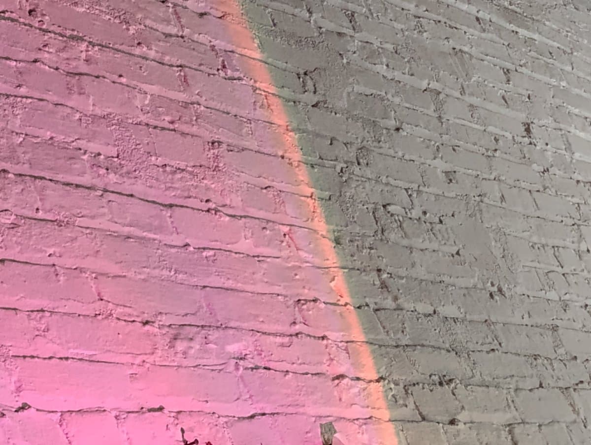 A close-up photograph of a brick wall painted in two distinct colors, with a pink section on the left and a grey section on the right, separated by a diagonal line.