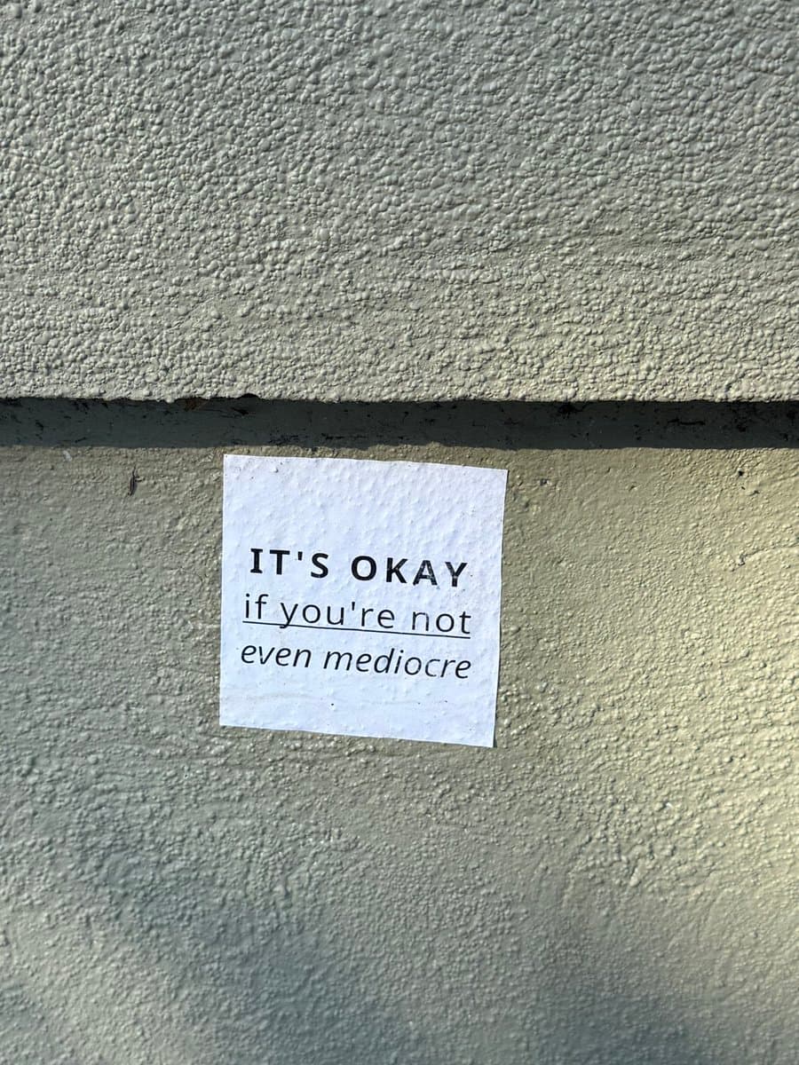 A small white piece of paper with printed text is taped to a textured, light-colored concrete wall. The paper contains a short, reassuring message.