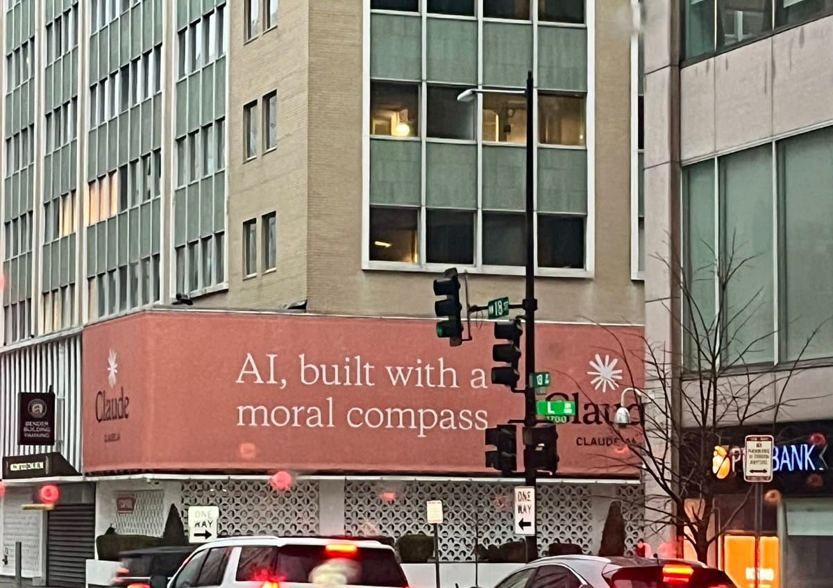 A street-level photograph of a city intersection featuring a large, reddish-brown billboard on the side of a building. The billboard displays the text "AI, built with a moral compass" alongside the Claude logo. Traffic lights, street signs, and the tops of cars are visible in the foreground.