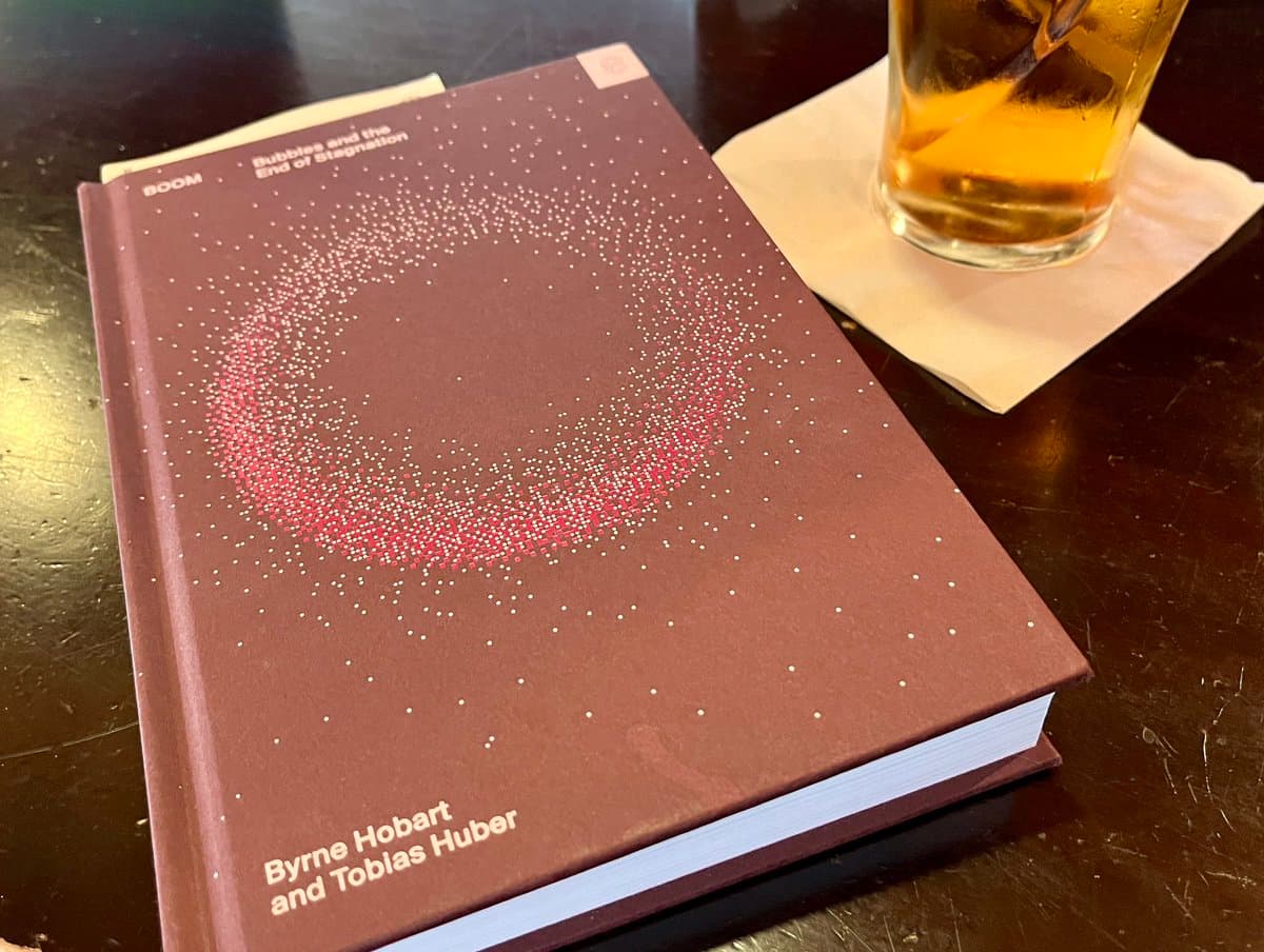A hardcover book titled "Bubbles and the End of Stagnation" by Byrne Hobart and Tobias Huber sits on a dark wooden table next to a glass of amber liquid on a napkin. The book cover is maroon with a dotted, crescent-shaped design in white and red.