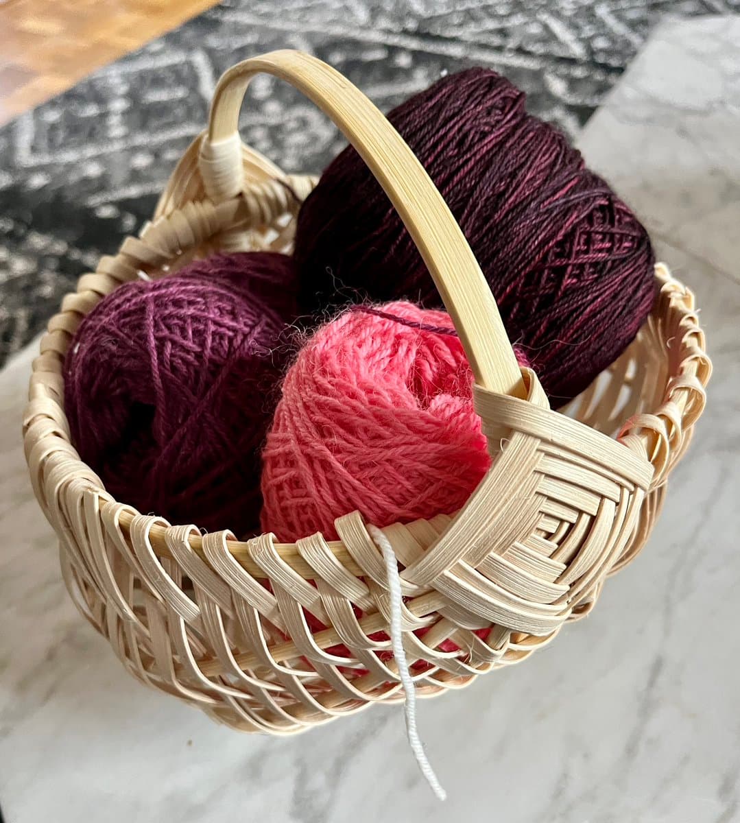 A small woven basket filled with three balls of yarn in shades of deep purple, maroon, and pink, resting on a marble surface. The basket has a light-colored handle and sits in front of a patterned fabric background.