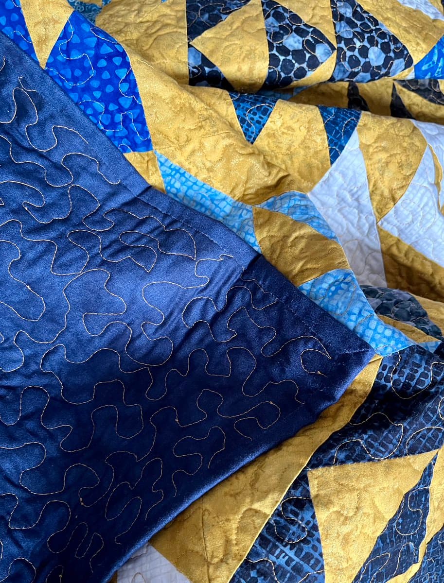 A close-up photograph of a handmade quilt featuring a patchwork of blue and gold fabrics with various patterns, including a solid blue section with decorative stitching.